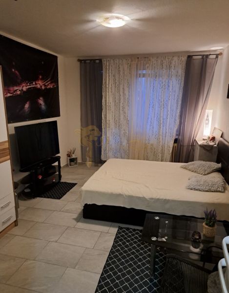 A studio apartment with gray-white walls, a bed, a television, and tiled flooring.