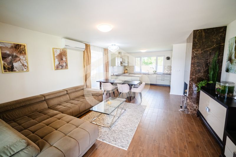 Living room in a family house with a kitchen, beige sofa, and wooden decor flooring.