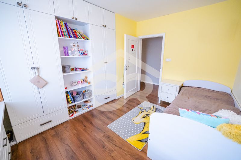 A children's room in a family house with a white wardrobe, a brown bed, and a wooden-patterned floor.