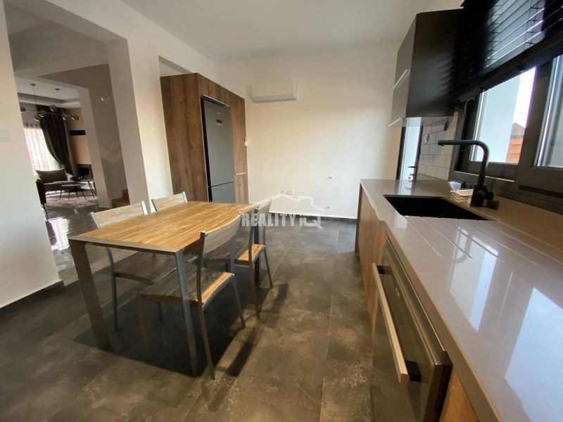 The kitchen of the family house shows a dining table, wooden elements, and a dark floor.