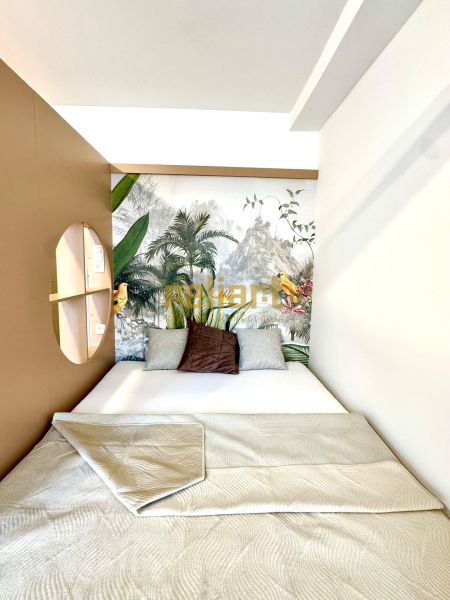 A bed with a patterned blanket in a studio apartment with tropical wallpaper and pillows.