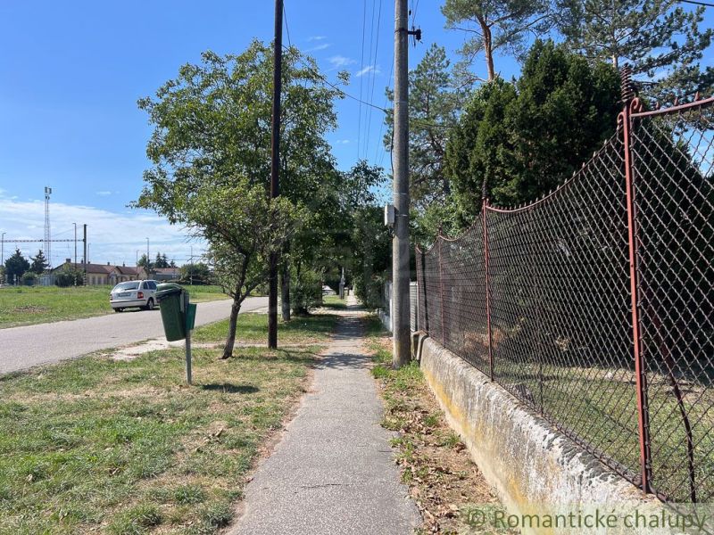 The sidewalk in Bernolákovo runs alongside a garden fence, lined with trees and grass.
