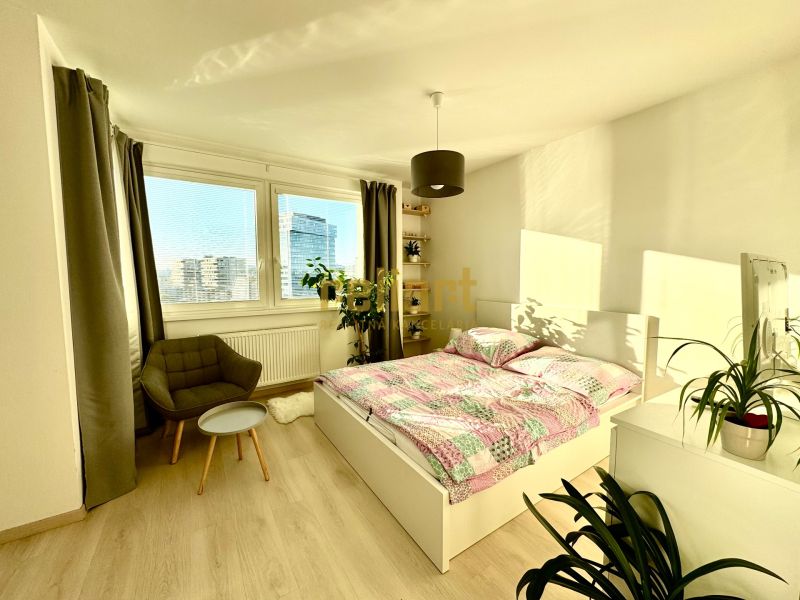 A bright bedroom with a bed, chair, and wood-decorated floor in a studio apartment.