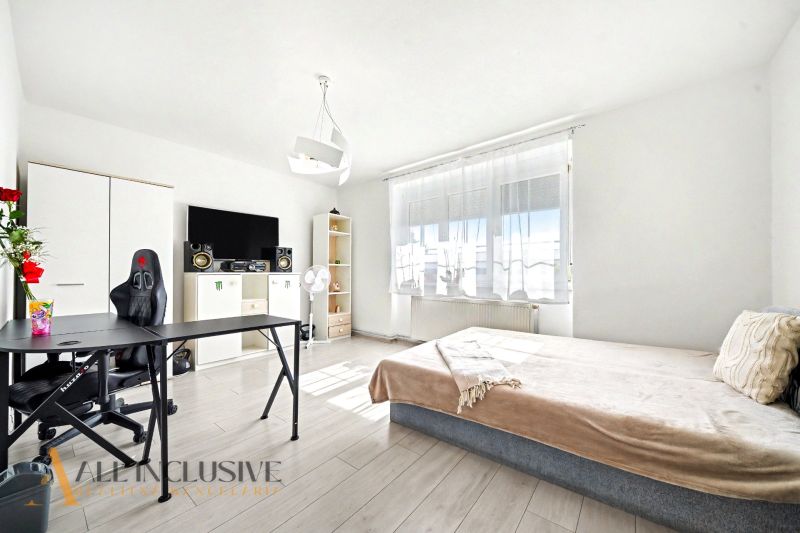 A bright room in a family house with a bed, a table, and a wooden-patterned floor.