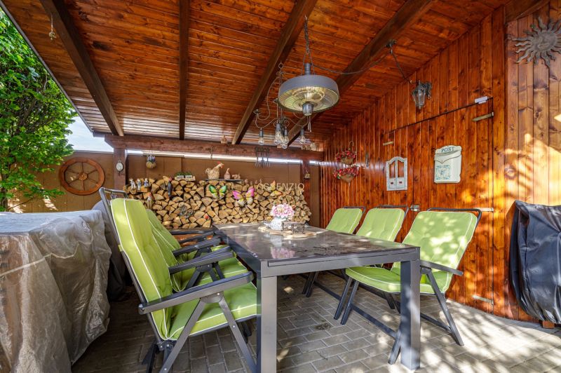 A wooden gazebo with a stone floor and green chairs by the family house.