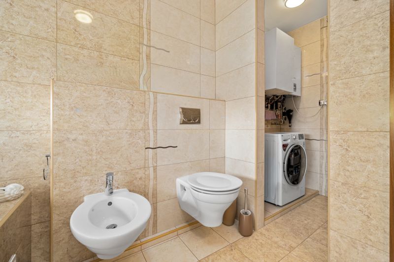 Bathroom with a toilet, bidet, and washing machine in a family house. Cream-colored tiles and flooring.