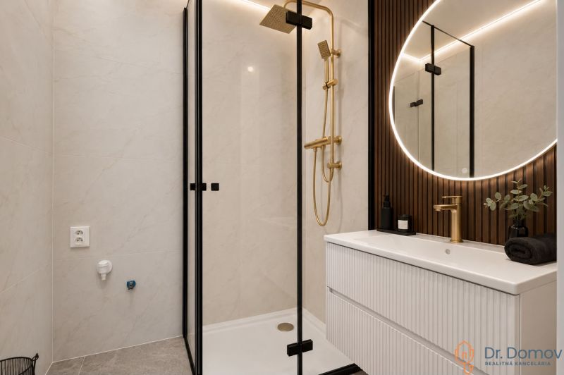 A bathroom in a 3-room apartment with an elegant shower, gold details, and a lighted mirror.