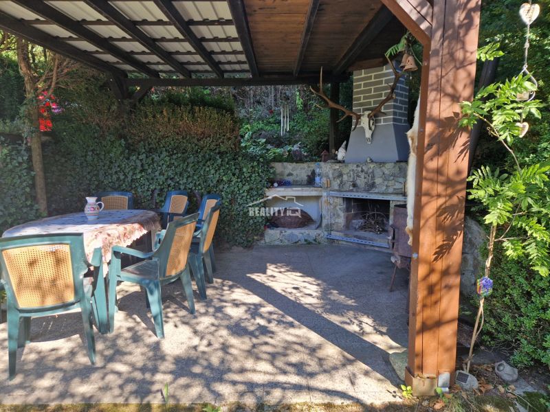 A covered terrace with a fireplace next to a cottage near Žilina, surrounded by greenery and seating.