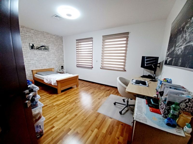 A room in a family house with a bed, a desk, and a wooden-patterned floor.