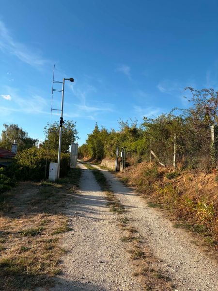 A reinforced dirt road leading to a cottage in Banka, lined with bushes and a fence.