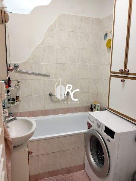A bathroom in a studio apartment with a bathtub and a washing machine, tiled with light tiles.