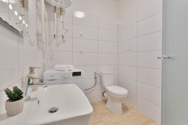 A bathroom in a studio apartment with a washing machine, sink, toilet, and shower, wooden decor flooring.