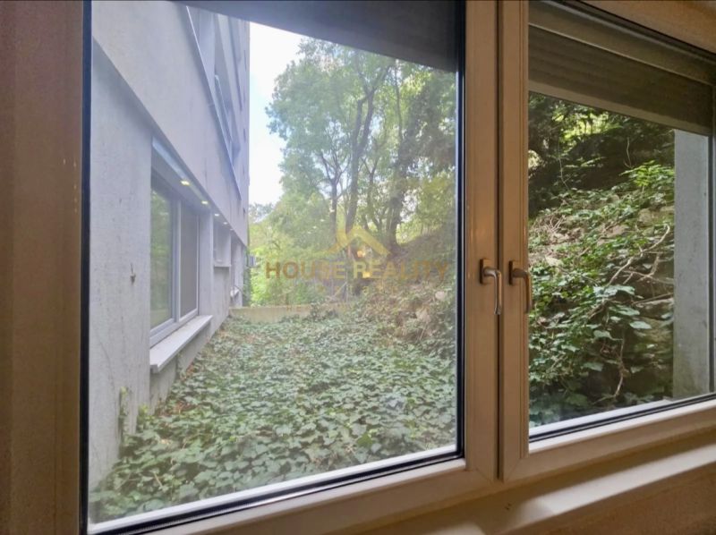 The view from the window of a one-room apartment onto greenery and an adjacent building.