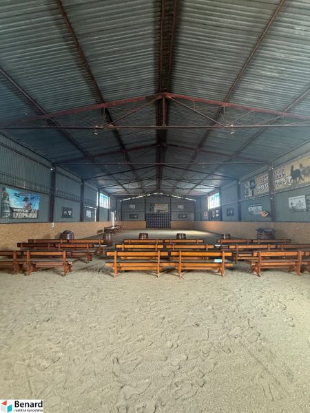 The interior of a building with wooden benches and a sandy floor, designed for events.