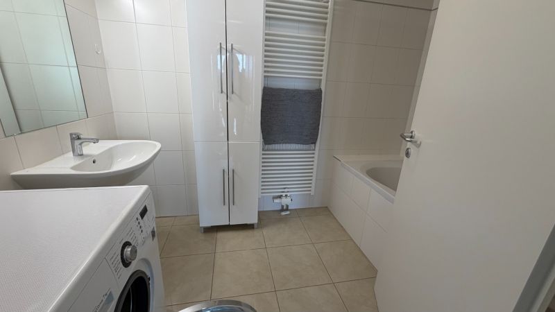 A bathroom in a 3-room apartment with a washing machine, sink, and bathtub on light tiles.