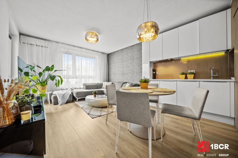 Living room in a two-room apartment with a wood-patterned floor, light furniture, and plants.