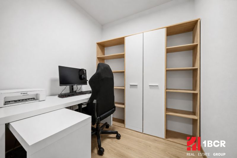 A workspace with a computer, printer, and shelves in a 2-room apartment with a wooden decor floor.