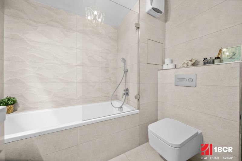 Bathroom with a bathtub and toilet in a 2-room apartment with light tiles.