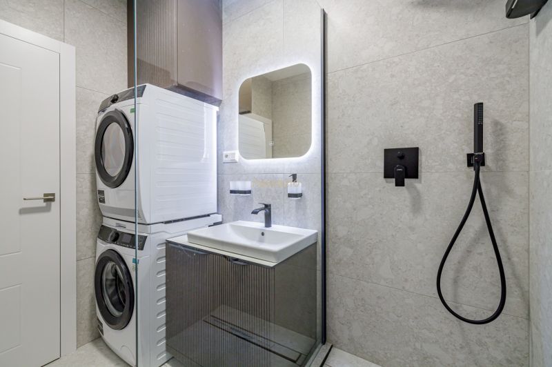 A bathroom with a washing machine, dryer, sink, illuminated mirror, and shower in a 3-room apartment.