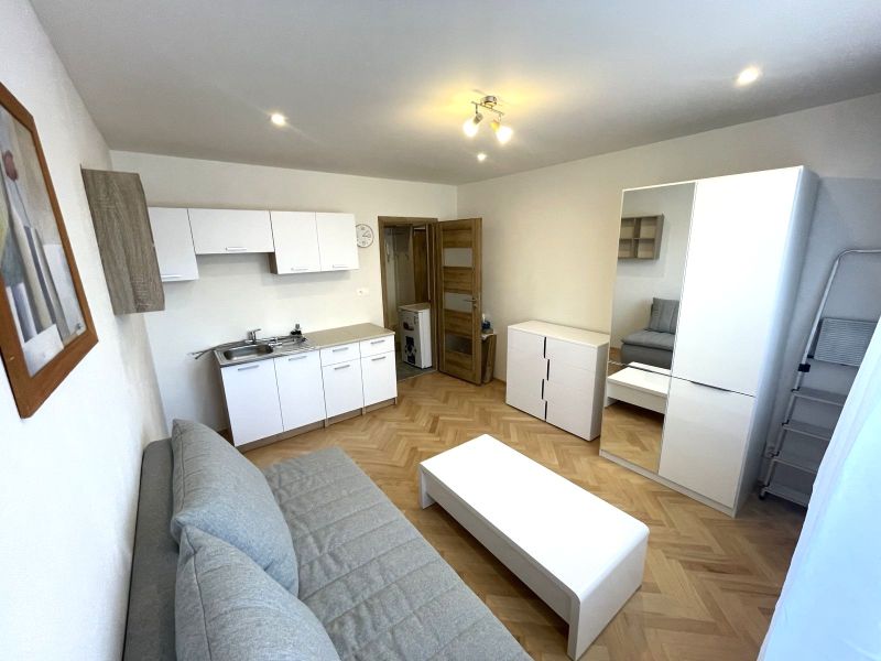 A studio apartment with a wooden decor floor, a kitchenette, and light-colored furniture.