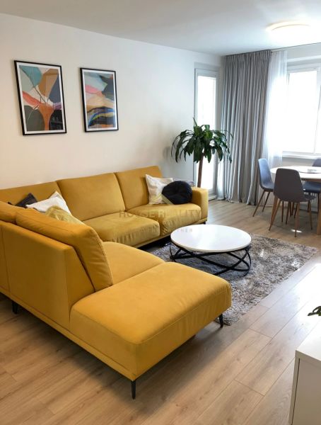 Living room of a 3-room apartment with a large yellow couch, wooden decor flooring, and a plant.