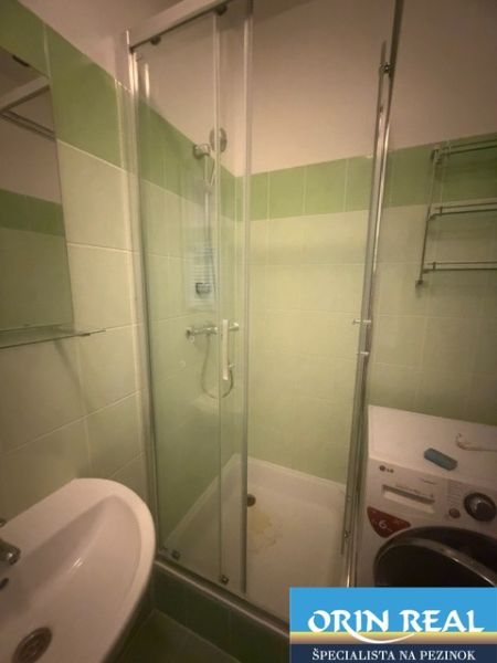 A bathroom in a studio apartment with a shower, washing machine, and sink with ceramic tiles.
