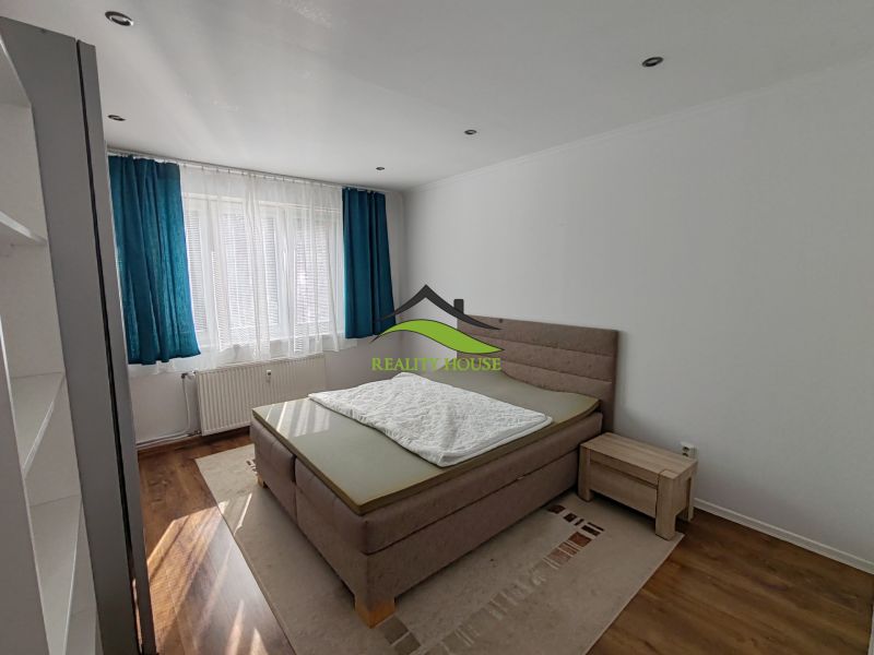 A bright bedroom in a three-room apartment with a wood-patterned floor and blue curtains.