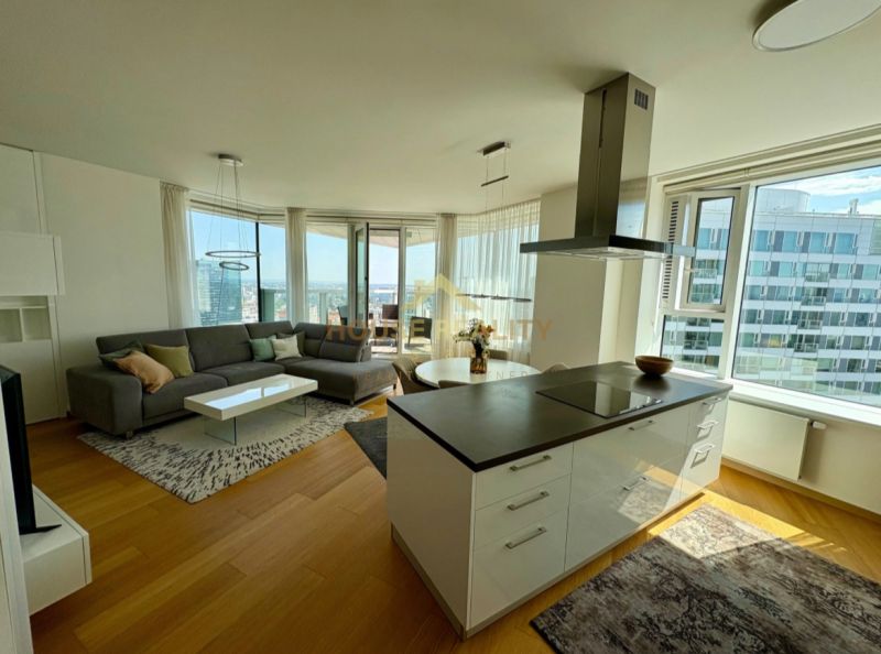 A bright living room with wood-patterned flooring in a three-room apartment, with a view from spacious windows.