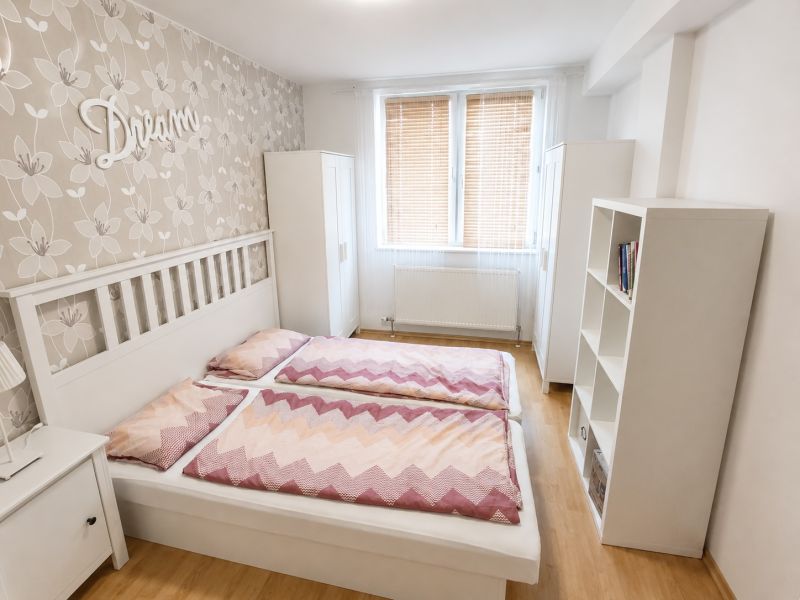 A bright bedroom with a double bed, floral wallpaper, and a wood-patterned floor.