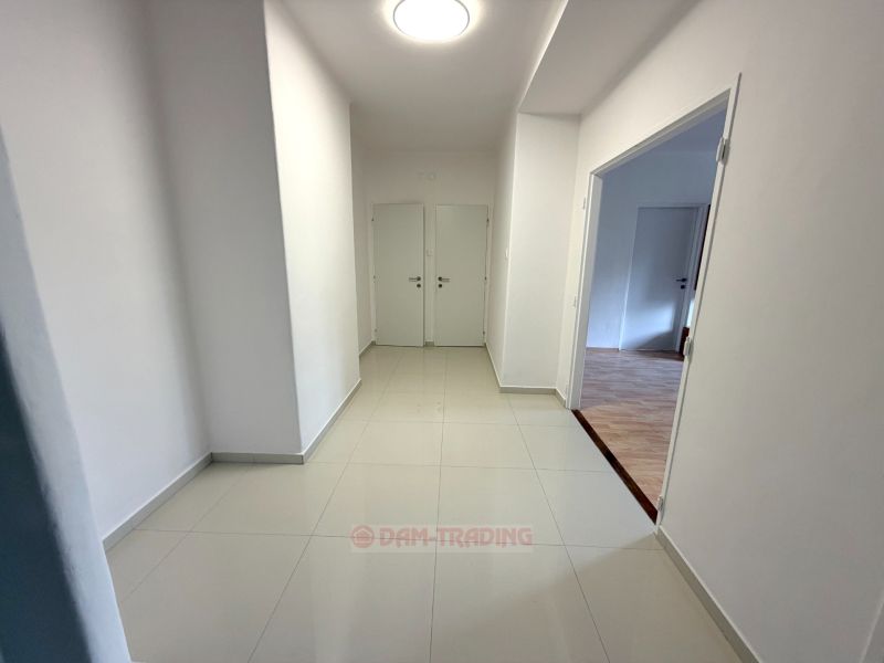 A corridor with light walls and tiles, entrance to a room with wood-patterned flooring.