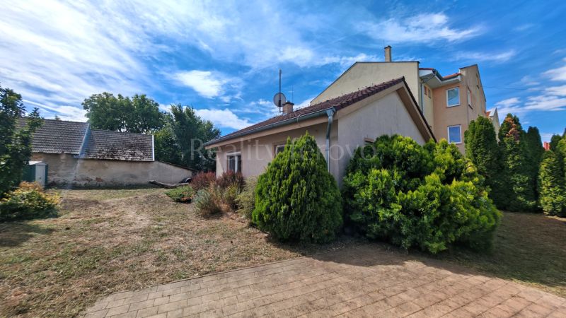 A family house in Matúškovo with a lawn, shrubs, and adjacent buildings under a blue sky.