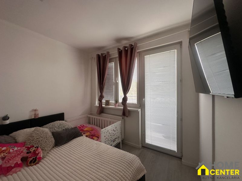 A bedroom in a 3-room apartment with a large window, curtains, and a TV on the wall.