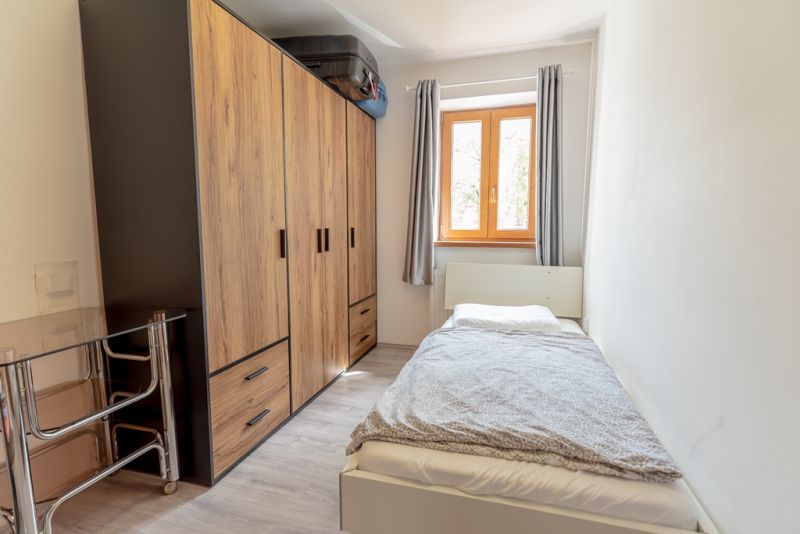 A small room in a family house with a bed, a built-in wardrobe, and a window with curtains.