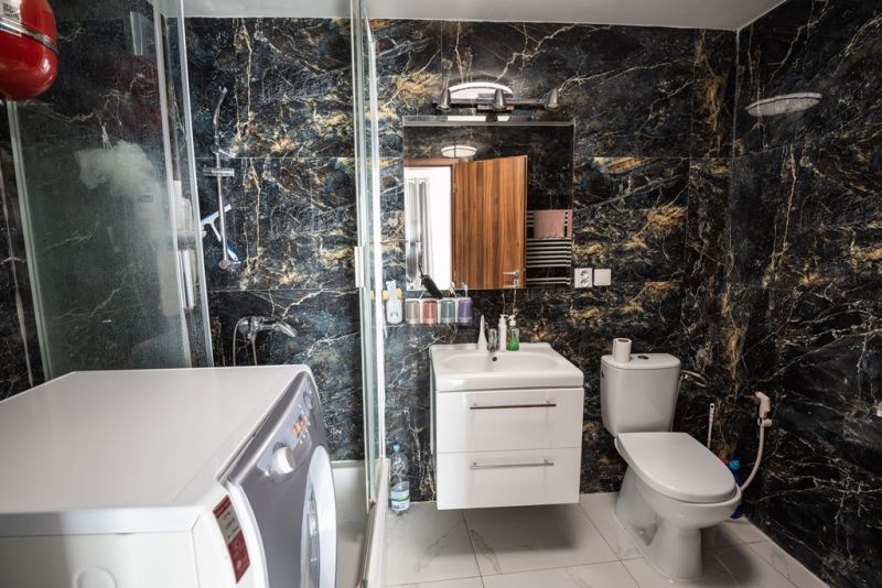 A bathroom in a family house with a washing machine, black marble walls, and a shower enclosure.