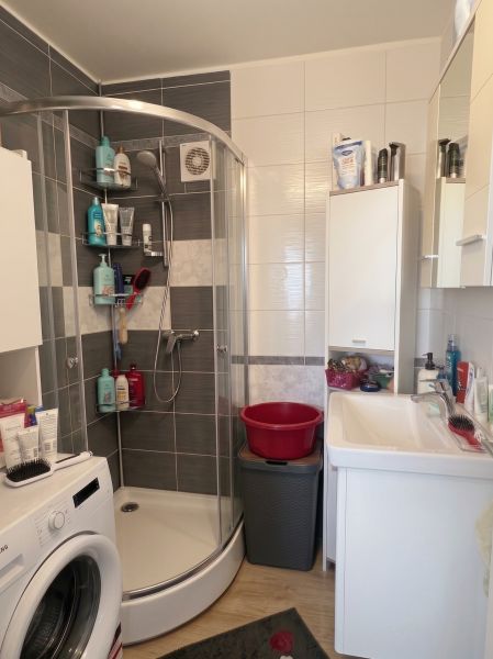 A bathroom with a shower, sink, washing machine, and shelf in a 3-room apartment.