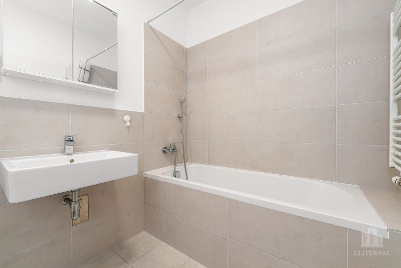 A bathroom with a bathtub and a sink in a two-room apartment. Neutral tiles on the walls.