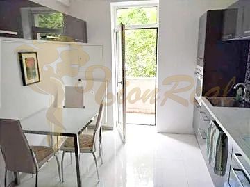 The kitchen of a two-room apartment with white tiles and a dining table by the window.