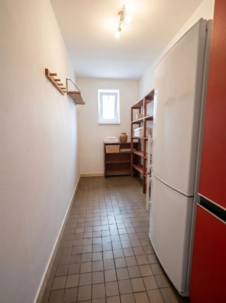 A narrow room in a family house with a refrigerator, shelves, a window, and a clothes hanger.