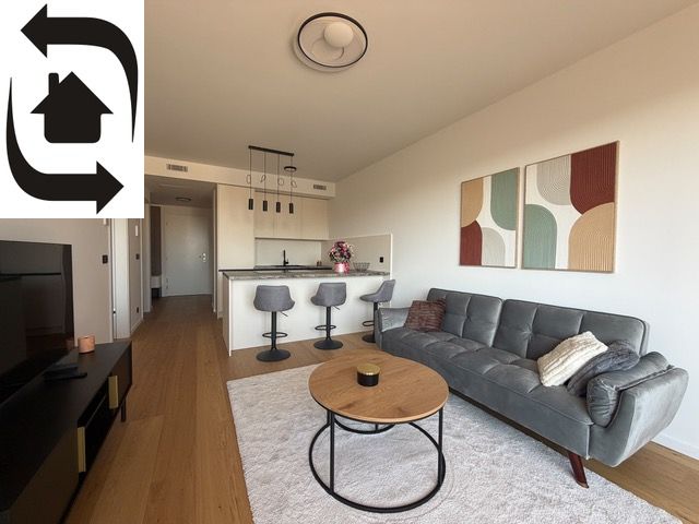 Living room in a two-room apartment with a wooden decor floor, a sofa, and a kitchenette.