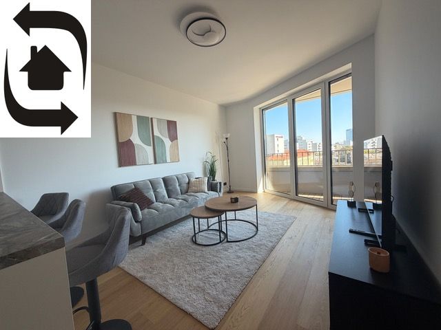 Living room in a two-room apartment with a sofa, balcony, and wooden decor flooring.
