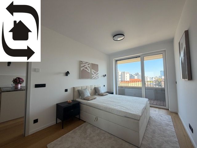 A bright bedroom in a two-room apartment with wood-patterned flooring and a balcony.