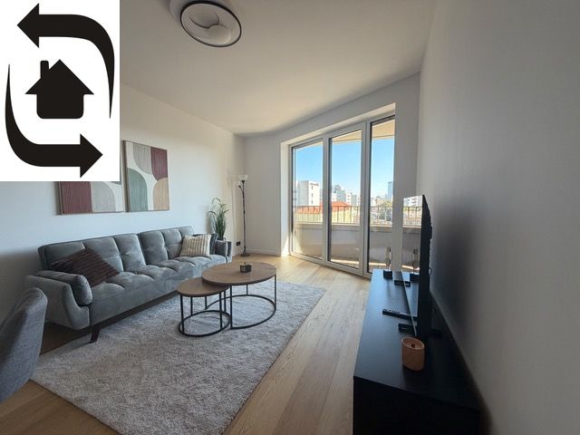 Living room in a two-room apartment with a wooden decor floor, large window, and sofa.