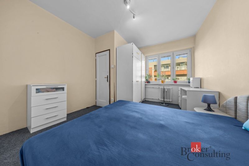 A room in a 3-room apartment with a bed, white cabinets, and windows with plants.