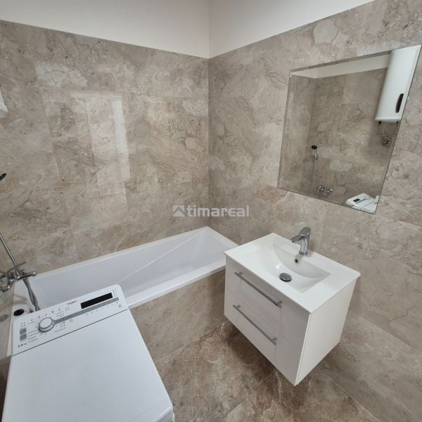 A bathroom in a studio apartment with a bathtub, sink, and washing machine; light stone tiles.