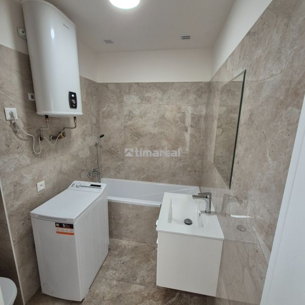 A bathroom with a bathtub, sink, and washing machine in a studio apartment, tiled with light tiles.