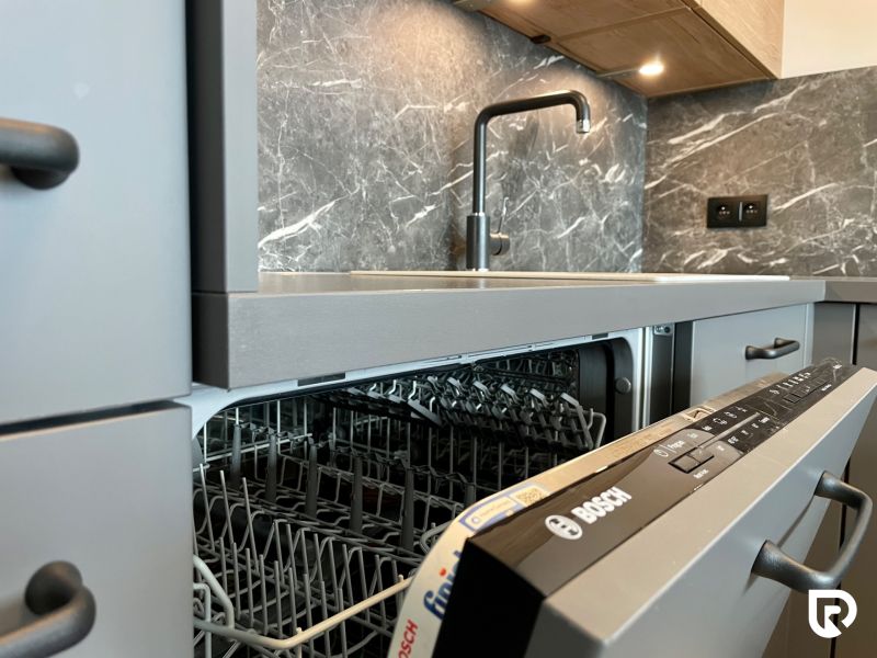 A kitchen unit in a 2-room apartment with an open dishwasher and gray cabinets.