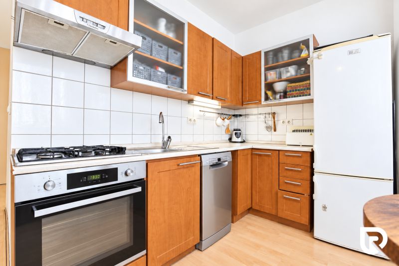 A kitchen with wooden decor, white tiles, a stove, and a stainless steel dishwasher in a three-room apartment.