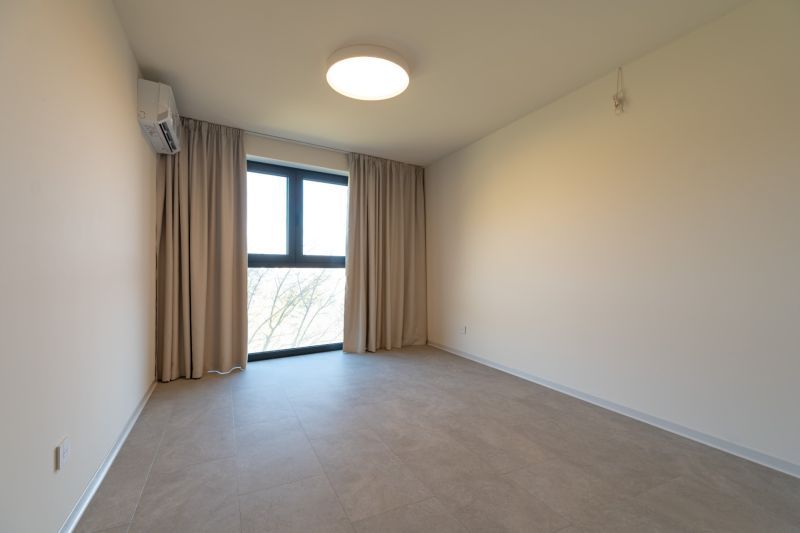 A room in a 4-room apartment with white walls, a large window, and air conditioning.