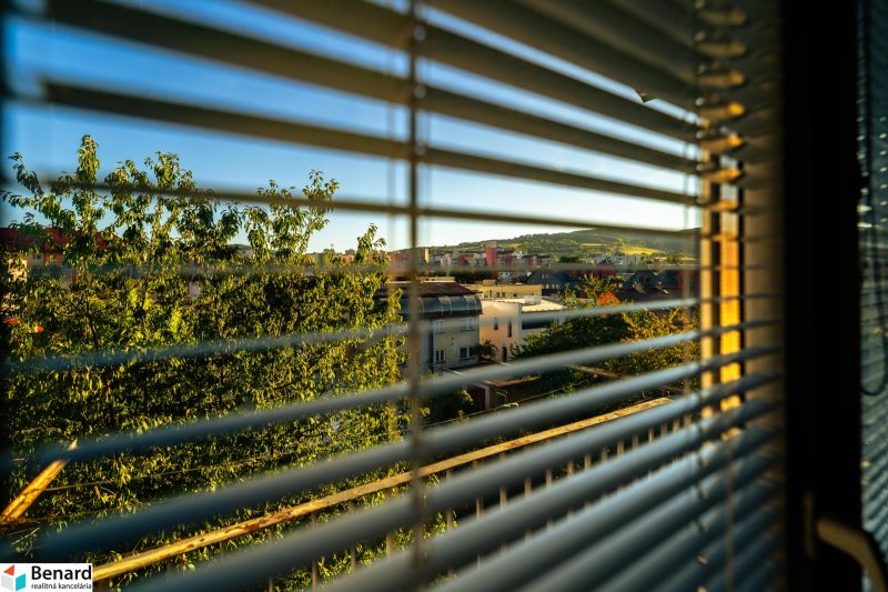 A view through the blinds of Bardejov from Fučíkova Street, overlooking greenery and buildings.