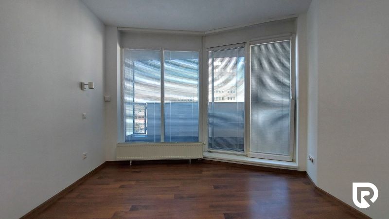 Living room of a two-room apartment with wood-patterned flooring and large windows with blinds.
